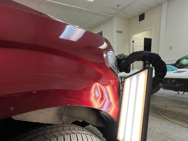 A dent repair light held against the front fender of a red SUV in the shop, a dent showing in the reflection