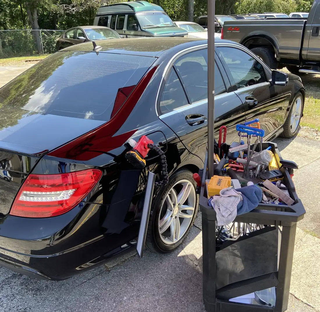 A cart of paintless dent repair rods, tabs and tools parked beside a black Mercedes sedan, a light set up on the rear quarter panel