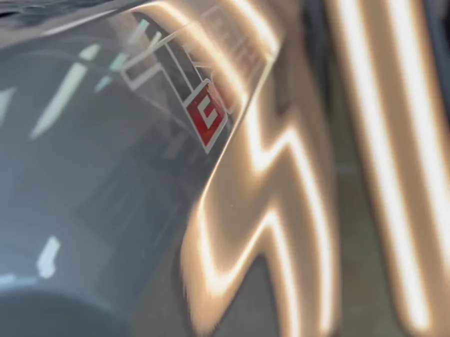 Two bright light strips reflected in a grey car's body line beside a red badge
