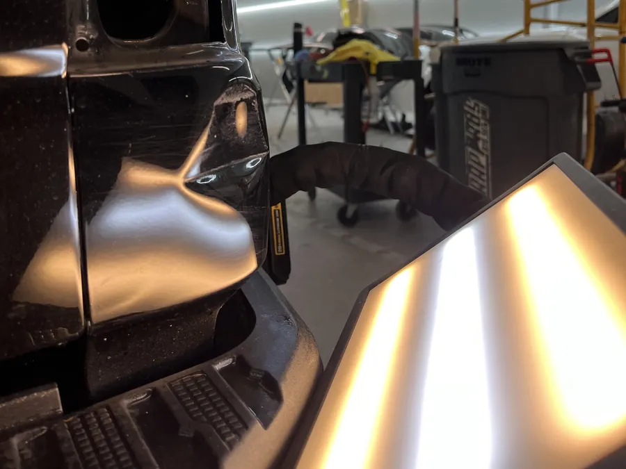 A light board set up beside a dark vehicle's bed side in the shop, its reflection running across the panel